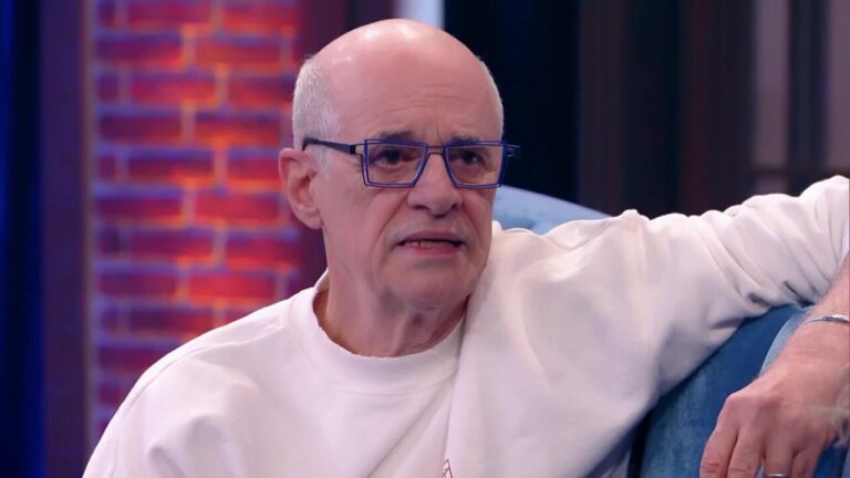 Homem calvo de óculos com armação azul, vestindo uma blusa branca e sentado em um sofá azul claro. Ele parece estar participando de uma entrevista ou conversa em um estúdio com iluminação colorida e parede de tijolos ao fundo.