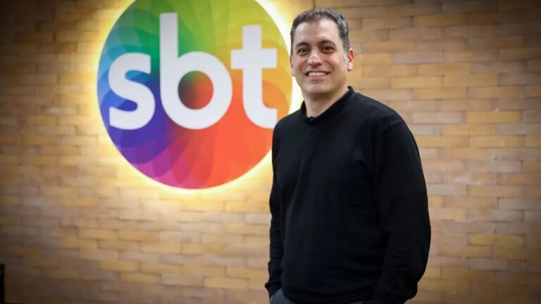 Marcos Kotait sorri em frente a uma parede de tijolos iluminada com o logotipo colorido do SBT. Ele veste suéter preto e calça escura, transmitindo um ar descontraído em ambiente corporativo da emissora de televisão