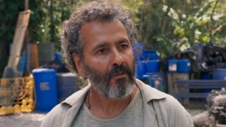 Homem de barba grisalha e expressão séria está ao ar livre, com tambores azuis e vegetação ao fundo, sugerindo um cenário rural ou industrial.