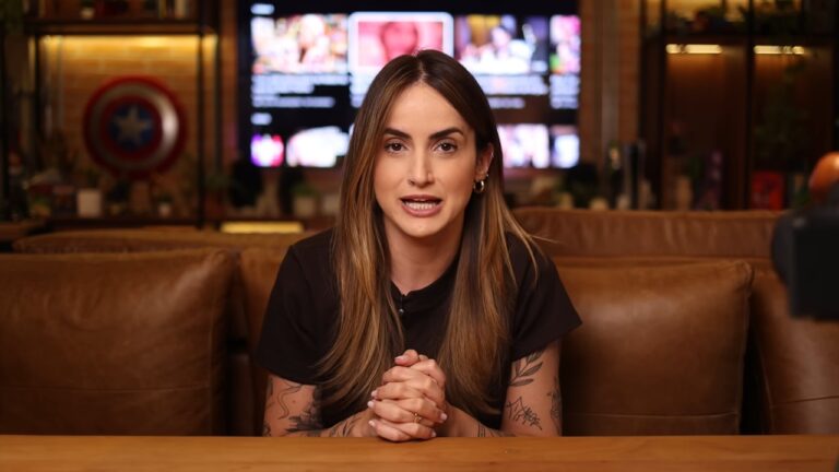 Mulher com tatuagens nos braços fala sentada à mesa em estúdio com sofá e tela desfocada ao fundo