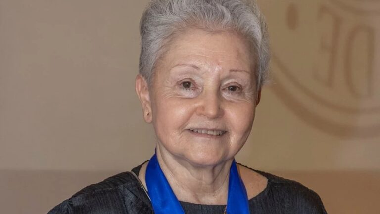 Mulher idosa sorri suavemente usando vestido escuro e medalha azul pendurada no pescoço, em ambiente interno de iluminação suave.