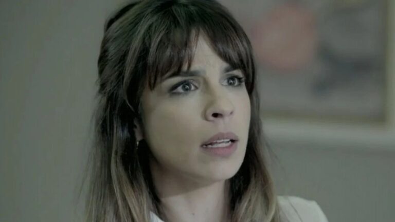 Mulher com expressão tensa e olhar fixo parece estar em uma cena de drama, com iluminação suave e fundo desfocado em ambiente interno.