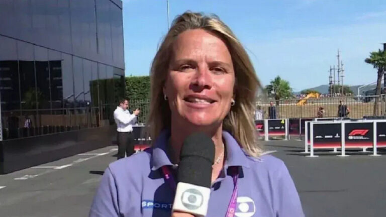 Mulher loira segura um microfone com o logotipo da TV Globo enquanto sorri para a câmera. Ela usa camisa polo roxa e crachá pendurado no pescoço. Ao fundo, há uma área externa ensolarada com pessoas caminhando, cercas metálicas e placas com o logotipo da Fórmula 1.