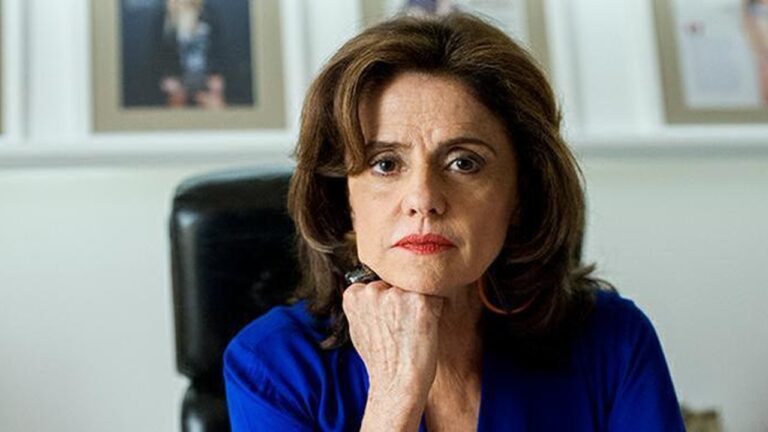 Mulher de expressão séria está apoiando o queixo na mão, vestindo uma blusa azul e batom vermelho. Ela está sentada em uma cadeira de escritório, com quadros desfocados ao fundo, transmitindo um ar de introspecção ou reflexão.