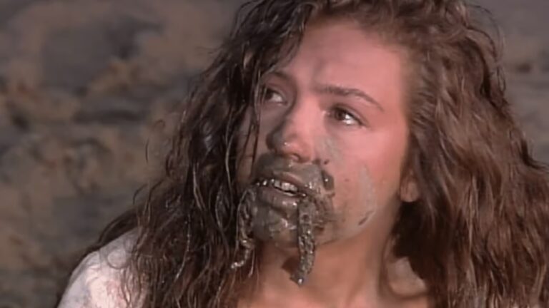 Mulher com o rosto e os cabelos sujos de lama olha para cima com expressão de tristeza ou desespero, em uma cena dramática ao ar livre.