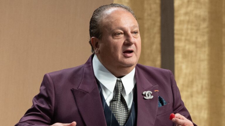 A imagem mostra Erick Jacquin usando um terno roxo elegante, camisa branca e gravata estampada. Ele também tem um broche da marca Chanel preso à lapela. A expressão é de fala, julgando a participação de Valesca Popozuda no MasterChef Celebridades, programa de televisão exibido pela Band