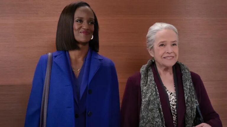 Duas mulheres sorriem lado a lado em ambiente interno com parede de madeira ao fundo, uma vestindo casaco azul e carregando bolsa no ombro, e a outra com cabelos grisalhos presos, usando roupa em tons de roxo e cachecol cinza.