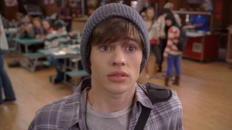 Jovem com expressão surpresa ou assustada, usando touca cinza, camisa xadrez e camiseta clara, com uma alça de mochila sobre o ombro. Ele está em um refeitório escolar, com mesas verdes e outros estudantes desfocados ao fundo, alguns em pé e outros sentados conversando.