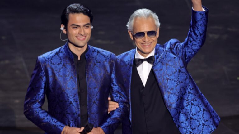 Dois homens no palco usam paletós azuis com padronagem floral brilhante, um deles segura um microfone e o outro acena sorrindo, vestindo óculos escuros, camisa branca e gravata borboleta preta, sob iluminação de espetáculo.