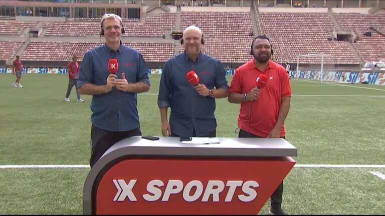 Mauro Beting, Marcelo do Ó e Leandro Guerreiro, da XSports, aparecem em campo de futebol durante transmissão ao vivo da final da Copinha. Eles seguram microfones vermelhos com o logotipo do canal e estão diante de uma bancada com a marca XSports em destaque. Ao fundo, vê-se parte das arquibancadas e jogadores realizando o aquecimento antes da partida