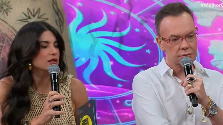 A imagem mostra Pâmela Lucciola e Felipeh Campos segurando microfones durante o programa Melhor da Noite. À esquerda, uma mulher de cabelos escuros e longos veste blusa bege rendada e usa brincos e anéis dourados. À direita, um homem de óculos vermelhos, camisa branca e acessórios chamativos, como anéis e pulseiras, também fala ao microfone. Ao fundo, há um painel colorido com símbolos astrológicos, sugerindo um quadro sobre horóscopos ou espiritualidade