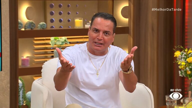Leo Dias, sentado em poltrona branca, fala de forma expressiva no estúdio do programa Melhor da Tarde, exibido ao vivo pela Band. Ele usa camiseta branca, colar dourado com crucifixo e gesticula com as mãos estendidas enquanto se dirige à câmera