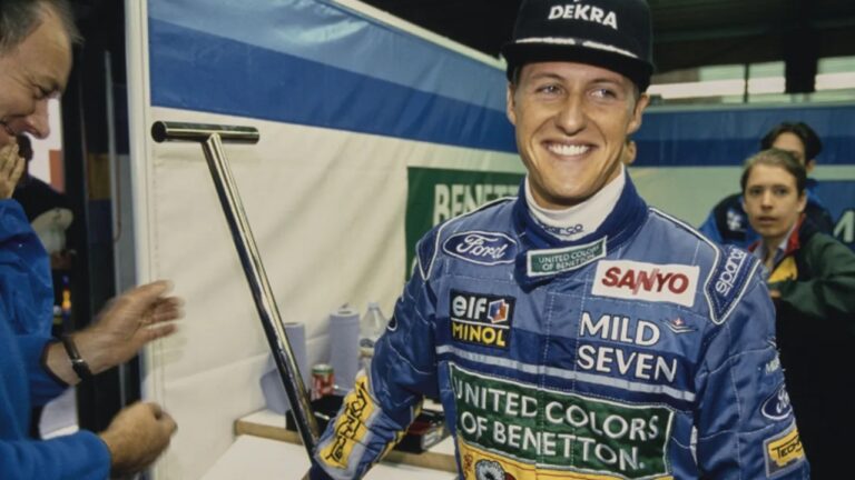 Piloto de automobilismo sorri nos boxes vestindo macacão azul com logotipos de patrocinadores e boné preto, em ambiente de equipe de corrida com outras pessoas ao fundo.