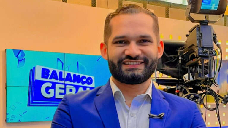 Miguel Anderson Costa sorri em selfie no estúdio do Balanço Geral com câmera de televisão e telão do programa ao fundo