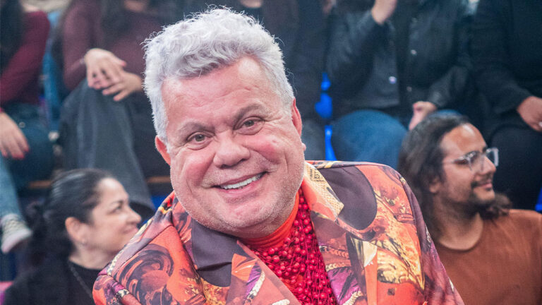 Milton Cunha sorri em estúdio de TV usando blazer estampado e colares vermelhos com plateia sentada ao fundo