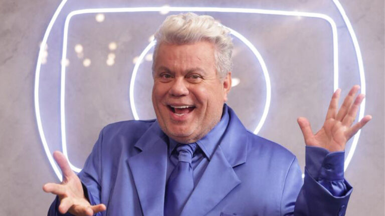 Milton Cunha sorridente de terno azul acetinado com as mãos espalmadas em frente ao logotipo da Globo em neon durante gravação da vinheta de fim de ano