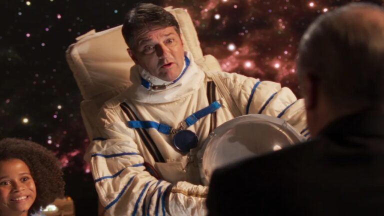Homem vestido com traje de astronauta segura o capacete enquanto conversa com outra pessoa em um cenário que simula o espaço, com estrelas e nebulosas ao fundo; uma criança observa sorridente ao lado