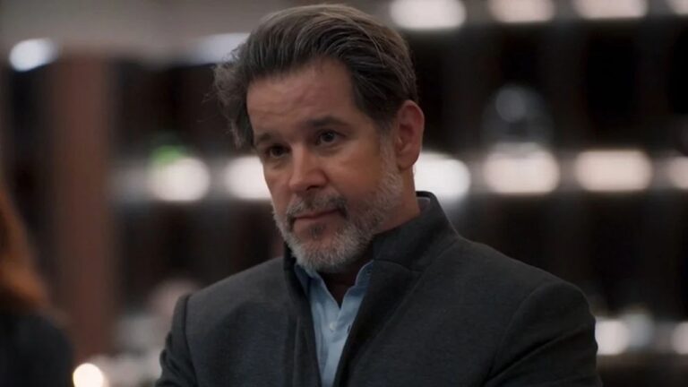 Homem de meia-idade com expressão séria está em um ambiente interno, vestindo camisa azul clara e paletó escuro, com prateleiras desfocadas ao fundo.