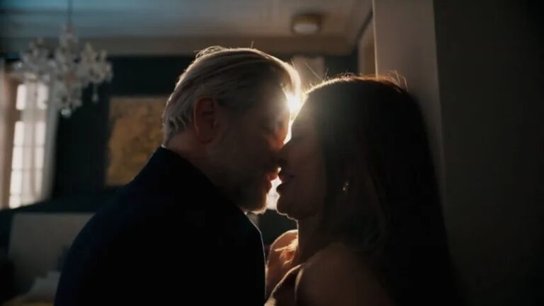 Homem de cabelos grisalhos e mulher se aproximam para beijo em ambiente interno com luz forte ao fundo criando silhuetas em cena dramática