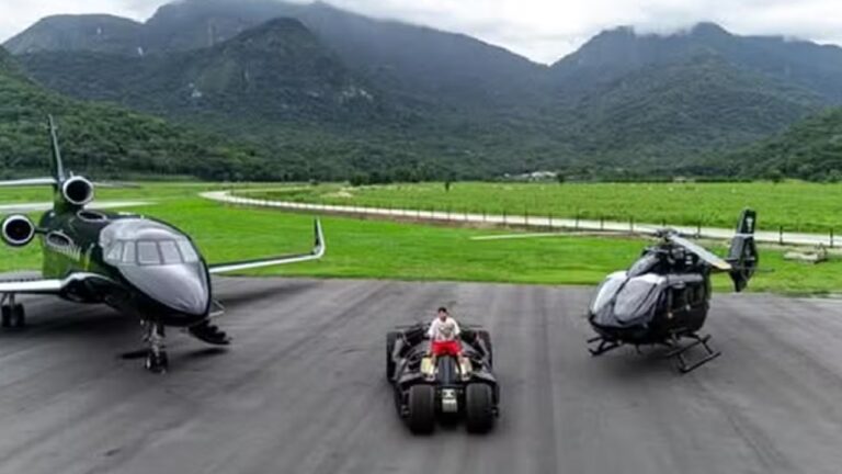 A imagem mostra uma pista de pouso com um jato particular preto à esquerda, um helicóptero preto à direita e, ao centro, um veículo estilizado semelhante ao Batmóvel. Uma pessoa está sentada sobre o veículo, vestindo roupas claras e vermelhas. Ao fundo, há uma paisagem montanhosa coberta de vegetação, sob céu nublado.