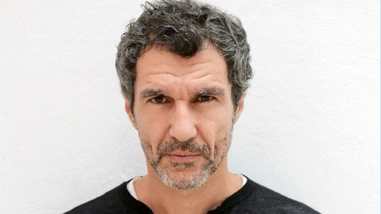 Nicola Siri encara a câmera com expressão séria usando camiseta preta e exibindo cabelos grisalhos curtos em fundo branco liso