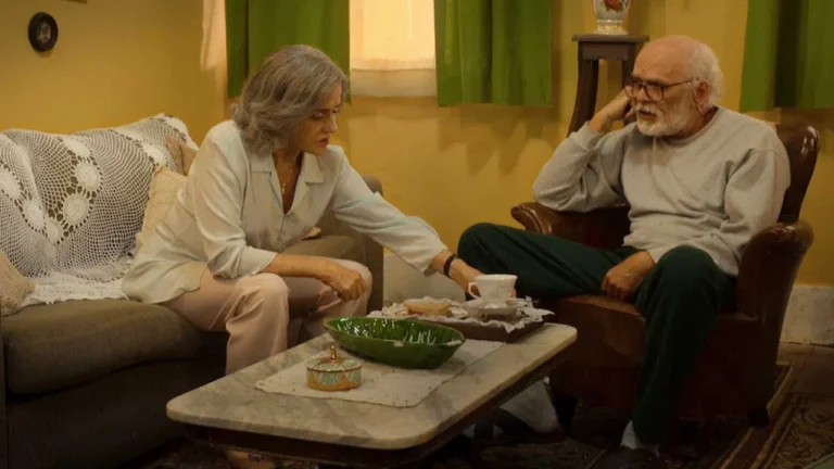 Cena em uma sala de estar simples e acolhedora, com luz amarelada. Um casal de idosos está em conversa: a mulher, sentada no sofá, se inclina para pegar algo sobre a mesa de centro, enquanto o homem, em uma poltrona, parece pensativo, apoiando a cabeça na mão. A mesa tem xícaras, pratos e uma tigela verde, e o ambiente transmite um clima doméstico e intimista.