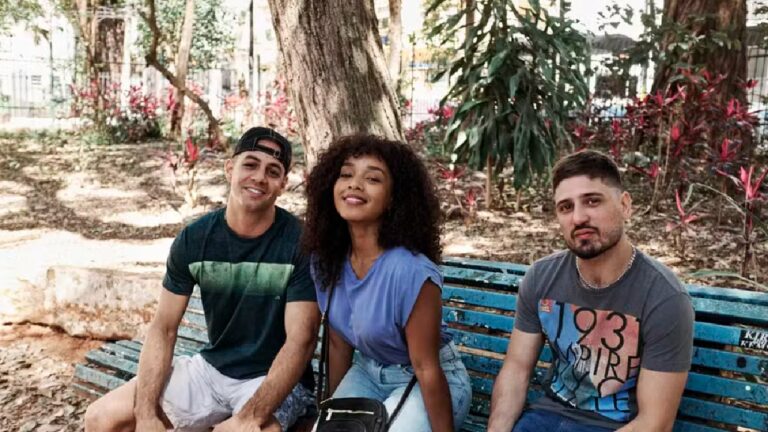 Três pessoas sentadas em um banco azul em parque arborizado posam sorrindo para a câmera em um dia ensolarado