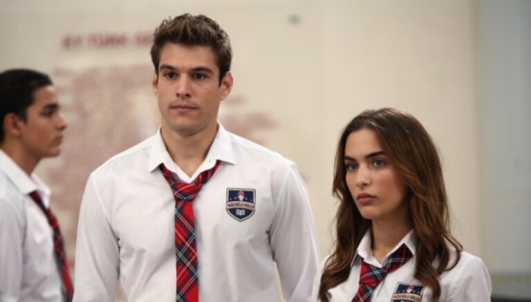 A imagem mostra dois jovens usando uniformes escolares — camisas brancas com gravatas xadrez vermelhas e brasões no peito. Eles estão lado a lado, com expressões sérias, em um ambiente que parece ser uma sala de aula ou colégio. Ao fundo, outro estudante aparece parcialmente desfocado, também usando o mesmo uniforme.