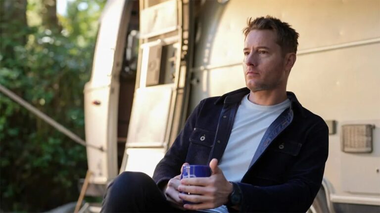 Homem de expressão séria está sentado ao ar livre segurando uma caneca azul, vestindo jaqueta escura e camiseta branca, com um trailer metálico e vegetação desfocada ao fundo sob luz natural do fim da tarde.