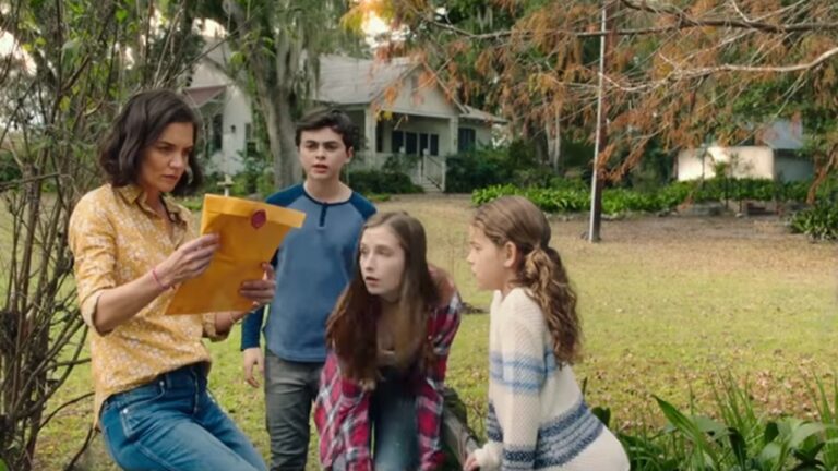 Mulher de camisa amarela segura um grande envelope pardo lacrado e o observa com atenção enquanto três crianças, duas meninas e um menino, olham curiosas ao seu redor. O grupo está em um gramado arborizado, com uma casa simples ao fundo e árvores com folhas alaranjadas indicando clima de outono.