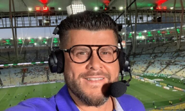 Comentarista esportivo usa headset e microfone em cabine de transmissão com estádio de futebol iluminado e arquibancadas ao fundo