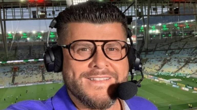 A imagem mostra um comentarista esportivo com fones de ouvido e microfone, provavelmente durante uma transmissão ao vivo de futebol. Ao fundo, é possível ver um estádio iluminado e o campo de jogo com jogadores se aquecendo.