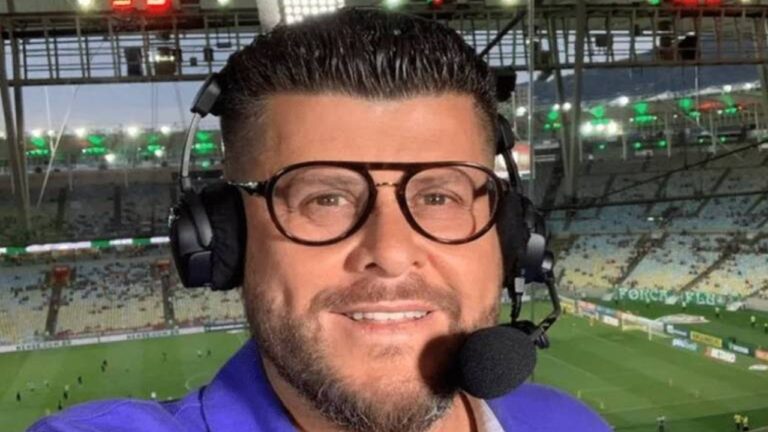 Homem usa fones de ouvido com microfone e óculos de armação grossa, sorrindo enquanto parece narrar ou comentar um evento esportivo. Ele está em um estádio de futebol iluminado, com parte do campo e arquibancadas visíveis ao fundo.