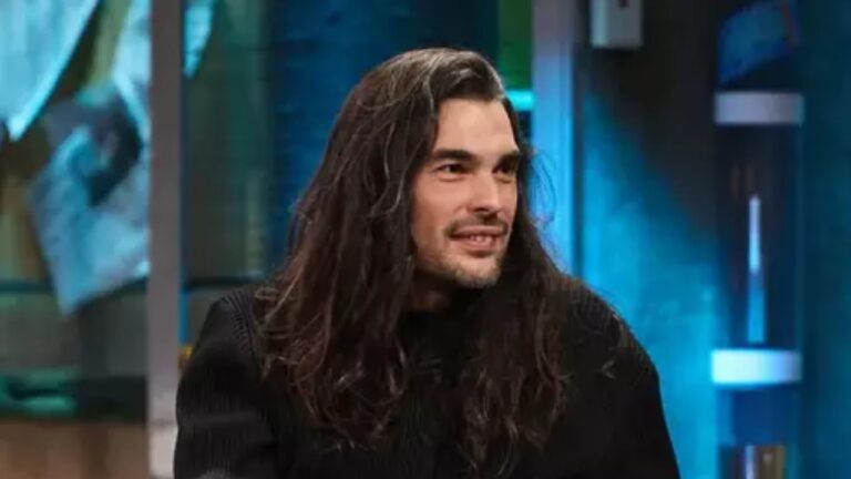 Homem de cabelos longos e suéter preto fala sorrindo durante entrevista em estúdio iluminado