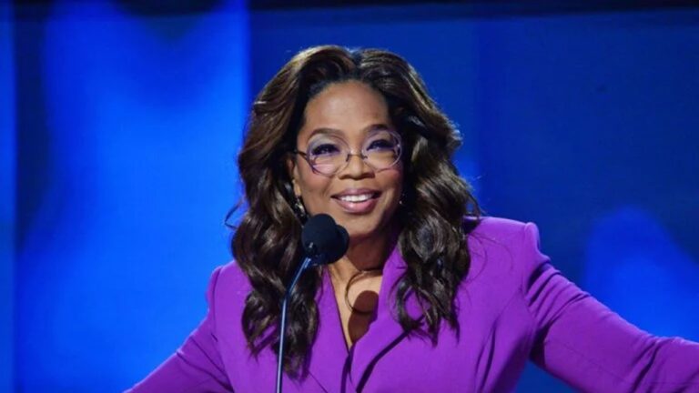 Oprah Winfrey sorri ao falar ao microfone em palco iluminado vestindo blazer roxo durante evento