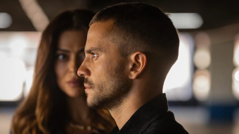 Homem de perfil encara à frente com mulher desfocada ao fundo em cena tensa de série policial