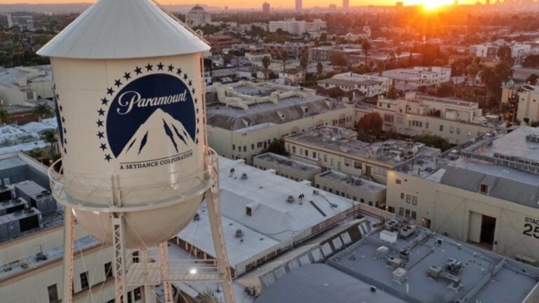 Vista aérea de um grande complexo de estúdios cinematográficos com destaque para a icônica caixa d’água branca da Paramount, que exibe o logotipo da montanha cercada por estrelas e a inscrição “A Skydance Corporation”. Ao fundo, o sol se põe atrás dos prédios da cidade, iluminando a paisagem urbana com tons alaranjados enquanto os galpões de produção e escritórios ocupam a maior parte da cena.