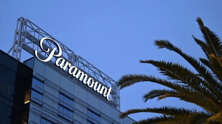 Texto Alternativo disse: A imagem mostra a fachada de um prédio moderno de vidro com o logotipo da Paramount no topo, escrito em letras brancas e cursivas. Em primeiro plano, há folhas de uma palmeira, e o céu azul claro ao fundo indica que a foto foi tirada durante o entardecer ou início da manhã.
