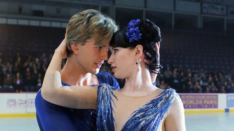 Casal de patinadores artísticos encara-se de perto em momento de emoção sobre o gelo, vestidos com figurinos azuis brilhantes diante do público