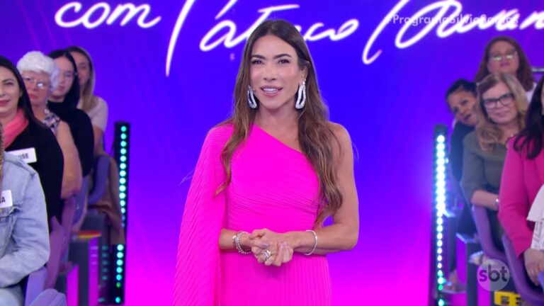Patricia Abravanel apresenta o Programa Silvio Santos em estúdio com iluminação roxa e rosa. Ela usa um vestido longo pink de um ombro só, brincos grandes prateados e pulseiras brilhantes. Ao fundo, o público acompanha sentado, e parte do letreiro do programa pode ser vista na parede com o logotipo do SBT no canto inferior direito