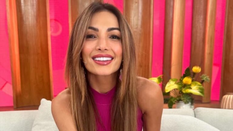 Mulher sorridente com blusa rosa apresenta programa em estúdio com fundo colorido e flores amarelas