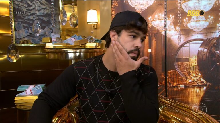 Paulo Augusto sentado em poltrona dourada no confessionário do BBB 26 aparece pensativo, com a mão no rosto. Ele usa boné preto virado para trás e suéter escuro com estampa geométrica. O ambiente é decorado com barras de ouro, notas de dinheiro e cristais pendurados, em cenário luxuoso