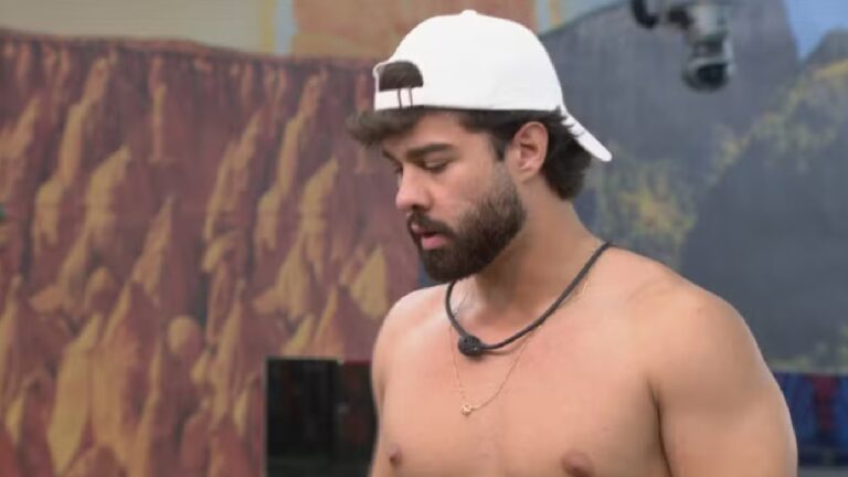 Homem barbudo, sem camisa e usando boné branco virado para trás, aparece com expressão concentrada em um ambiente externo decorado com painéis coloridos e câmeras visíveis, sugerindo a gravação de um reality show.