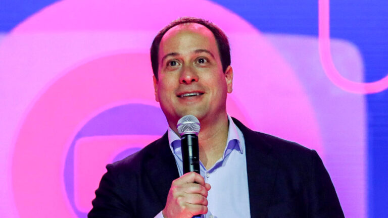 Paulo Marinho fala ao microfone vestindo terno azul marinho e camisa clara em frente a um telão com o logotipo da Globo ao fundo