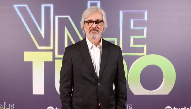 Paulo Silvestrini de terno preto e camisa branca posa diante de painel com logotipo da novela Vale Tudo em evento de lançamento