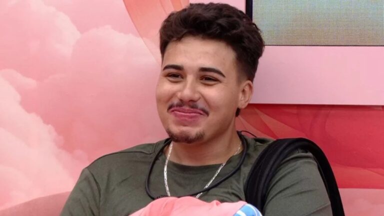 Homem jovem sorri sentado em ambiente interno com fundo em tons de rosa usando camiseta verde e colar visível no pescoço