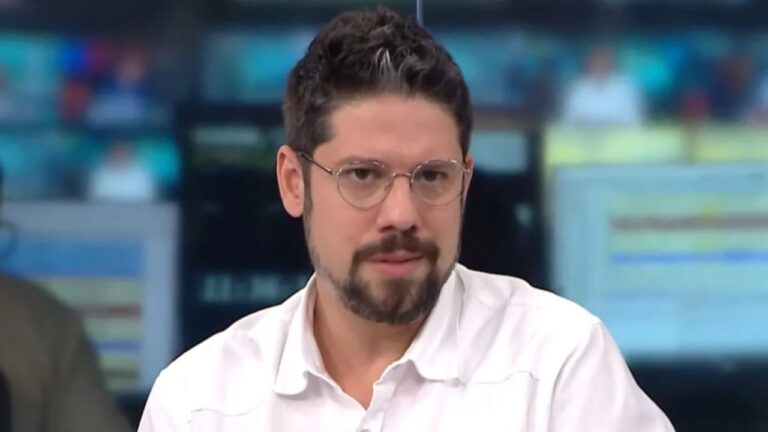 Homem de óculos e camisa branca fala em estúdio de jornal com telas desfocadas ao fundo