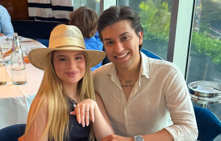 Um casal posa sorridente em um restaurante. A mulher, usando chapéu de palha e blusa preta, mostra a mão com um anel em destaque, enquanto o homem ao lado veste camisa clara e exibe uma tatuagem no peito. Ao fundo, há mesas postas com garrafas de água e pessoas sentadas, e o ambiente é iluminado por luz natural vinda de grandes janelas.