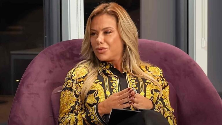 Mulher sentada em poltrona roxa conversa durante uma entrevista ou programa de televisão em ambiente interno bem iluminado. Ela usa blazer preto e amarelo com estampa dourada de arabescos, tem cabelos loiros soltos e unhas pintadas de vermelho escuro, segurando um caderno nas mãos enquanto olha para o lado.