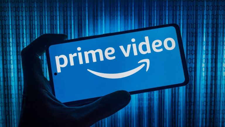 Mão segura smartphone exibindo o logotipo do serviço de streaming Prime Video em tela azul, com fundo digital iluminado em tons de azul e códigos binários.
